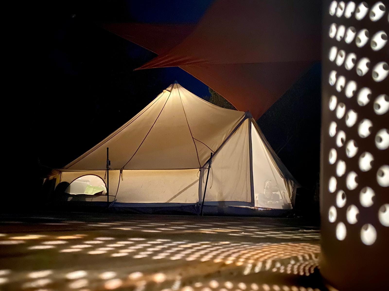 Cream bell tent lit from within at night, a lamp casting dots across the deck